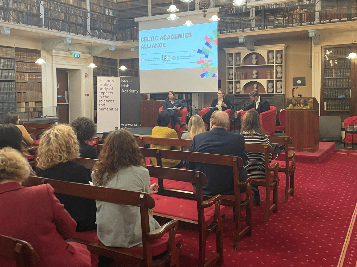 A meeting of the Celtic Academies Alliance at the Royal Irish Academy in Dublin. Three panellists sits in chairs in a panelled library faced by rows of attendees on upholstered wooden benches.