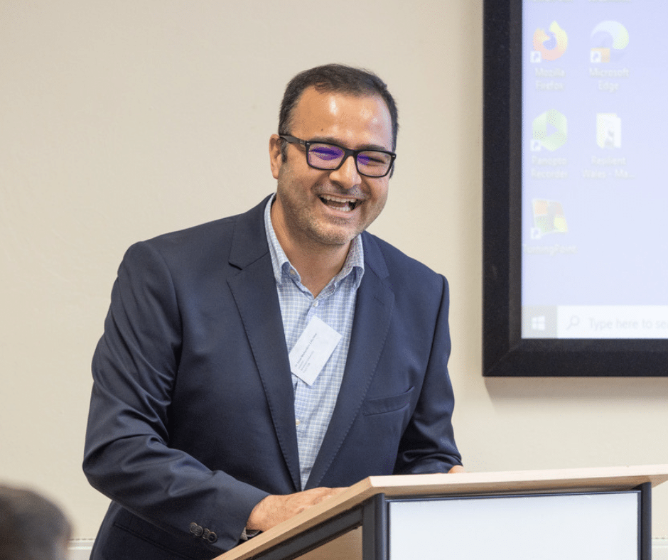 A man with a broad smile stands at a lecture with a large screen behind him.